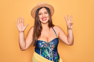 Young hispanic woman wearing summer hat and swimsuit over yellow background showing and pointing up with fingers number eight while smiling confident and happy.