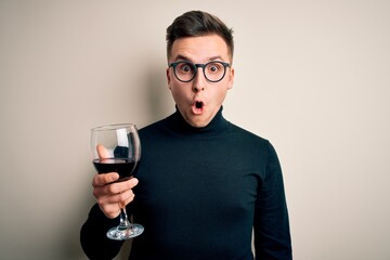 Young handsome caucasian man drinking an alcoholic glass of red wine over isolated background scared in shock with a surprise face, afraid and excited with fear expression