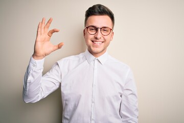 Young handsome business mas wearing glasses and elegant shirt over isolated background smiling and confident gesturing with hand doing small size sign with fingers looking and the camera. Measure