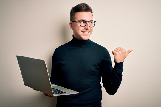 Young handsome caucasian business man wearing glasses using computer laptop pointing and showing with thumb up to the side with happy face smiling