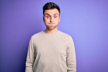 Young handsome caucasian man wearing casual sweater over purple isolated background puffing cheeks with funny face. Mouth inflated with air, crazy expression.