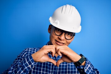 Young handsome engineer latin man wearing safety helmet over isolated blue background smiling in love showing heart symbol and shape with hands. Romantic concept.