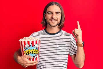 Young handsome man holding popcorn smiling with an idea or question pointing finger with happy face, number one