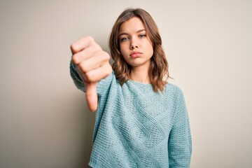 Young blonde girl wearing casual blue winter sweater over isolated background looking unhappy and angry showing rejection and negative with thumbs down gesture. Bad expression.