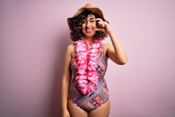 Young beautiful arab woman on vacation wearing swimsuit and hawaiian lei flowers smiling and confident gesturing with hand doing small size sign with fingers looking and the camera. Measure concept.