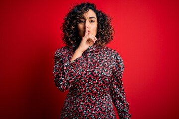 Young beautiful curly arab woman wearing casual floral dress standing over red background asking to be quiet with finger on lips. Silence and secret concept.