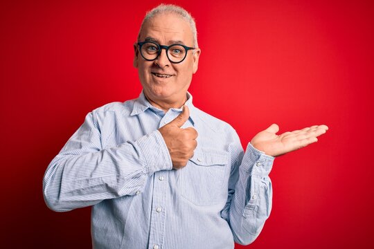Middle age handsome hoary man wearing casual striped shirt and glasses over red background Showing palm hand and doing ok gesture with thumbs up, smiling happy and cheerful