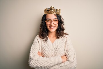 Young beautiful woman with curly hair wearing golden queen crown over white background happy face smiling with crossed arms looking at the camera. Positive person. © Krakenimages.com