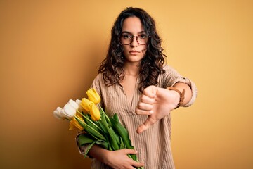 Young beautiful romantic woman with curly hair holding bouquet of yellow tulips looking unhappy and angry showing rejection and negative with thumbs down gesture. Bad expression.