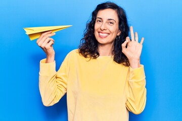 Young beautiful hispanic woman holding paper airplane doing ok sign with fingers, smiling friendly...