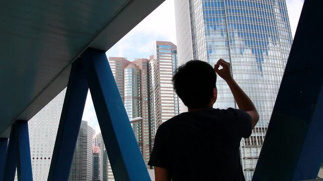 Young asian man in the footbridge looking through his hands up in front of a building seen from his back in Hong Kong International Finance Centre