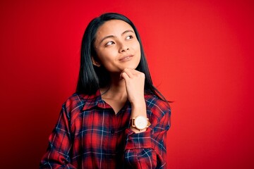 Fototapeta premium Young beautiful chinese woman wearing casual shirt over isolated red background with hand on chin thinking about question, pensive expression. Smiling and thoughtful face. Doubt concept.