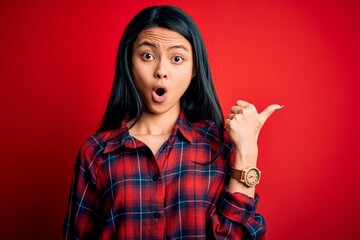 Young beautiful chinese woman wearing casual shirt over isolated red background Surprised pointing with hand finger to the side, open mouth amazed expression.