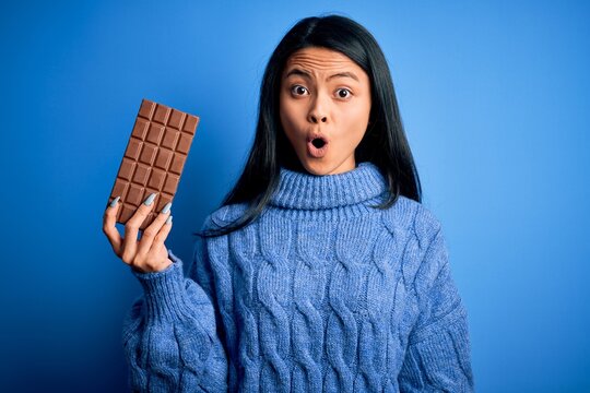Young beautiful chinese woman holding chocolate bar over isolated blue background scared in shock with a surprise face, afraid and excited with fear expression