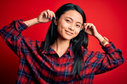 Young beautiful chinese woman wearing casual shirt over isolated red background Smiling pulling ears with fingers, funny gesture. Audition problem