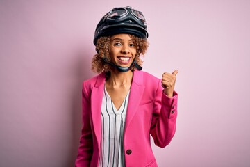 African american motorcyclist woman with curly hair wearing moto helmet over pink background smiling with happy face looking and pointing to the side with thumb up.