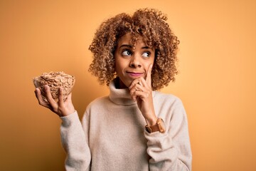 Young african american curly woman holding bowl with healthy cornflakes cereals serious face thinking about question, very confused idea