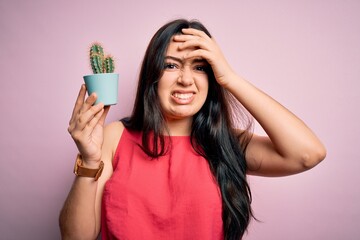Young brunette woman holding succulent cactus plant over pink background stressed with hand on...