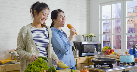 Two cheerful friends cook together and singing with carrot as microphone. asian girl sisters having fun dancing in kitchen with fresh vegetables. happy women laughing joyfully while prepare meal.