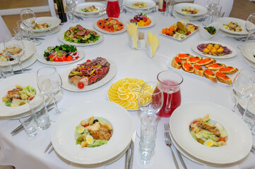 Banquet table with portioned caesar salads, meat and vegetable cuts, cold snacks and drinks. White tablecloth and plates. Red caviar sandwiches, roast beef, cheese assortment.