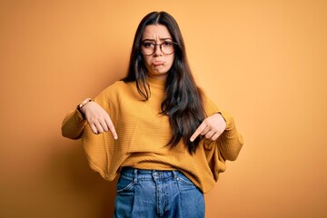 Young brunette woman wearing glasses and casual sweater over yellow isolated background Pointing down looking sad and upset, indicating direction with fingers, unhappy and depressed.