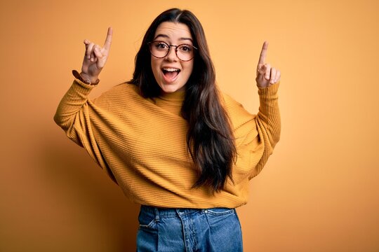 Young brunette woman wearing glasses and casual sweater over yellow isolated background smiling amazed and surprised and pointing up with fingers and raised arms.