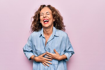 Middle age beautiful woman wearing casual denim shirt standing over pink background smiling and laughing hard out loud because funny crazy joke with hands on body. - obrazy, fototapety, plakaty