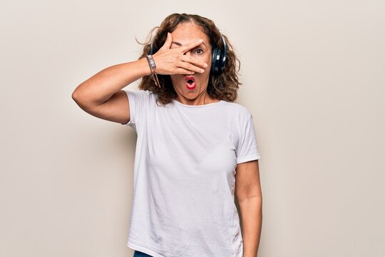 Middle age beautiful woman listening to music using headphones over white background peeking in shock covering face and eyes with hand, looking through fingers afraid