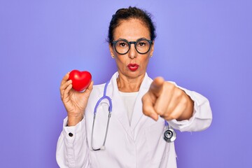 Middle age senior cardiologist doctor woman holding red heart over purple background pointing with finger to the camera and to you, hand sign, positive and confident gesture from the front