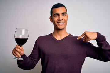 Handsome african american sommelier man tasting glass of red wine over white background with surprise face pointing finger to himself