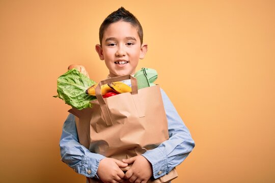 Adorable toddler holding paper bag with food standing over isolated yellow background