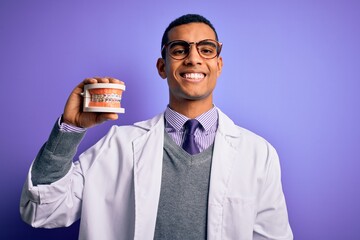 Young handsome african american dentist man holding denture teeth with dental braces with a happy face standing and smiling with a confident smile showing teeth