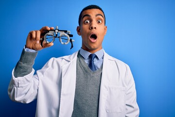 Handsome african american optical man holding optometry glasses over blue background scared in shock with a surprise face, afraid and excited with fear expression