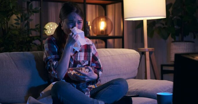 Sad woman crying and eating popcorn while watching movie at night