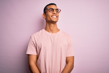 Handsome african american man wearing casual t-shirt and glasses over pink background looking away to side with smile on face, natural expression. Laughing confident.