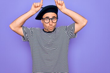 Naklejka premium Handsome man with blue eyes wearing striped t-shirt and french beret over purple background doing funny gesture with finger over head as bull horns