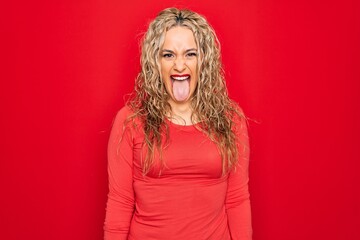 Young beautiful blonde woman wearing red casual t-shirt standing over isolated background sticking tongue out happy with funny expression. Emotion concept.