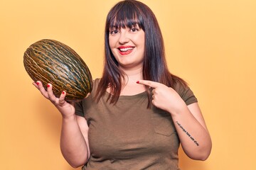 Young plus size woman holding melon smiling happy pointing with hand and finger