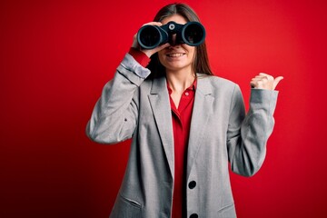 Young beautiful woman with blue eyes using binoculars over isolated red background pointing and showing with thumb up to the side with happy face smiling