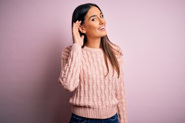 Fototapeta premium Young beautiful brunette woman wearing casual sweater over isolated pink background smiling with hand over ear listening an hearing to rumor or gossip. Deafness concept.