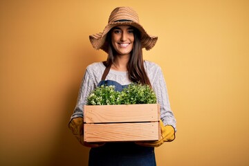 Young beautiful brunette gardener woman wearing apron and hat holding box with plants with a happy...