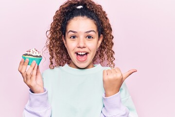 Beautiful kid girl with curly hair holding cupcake pointing thumb up to the side smiling happy with open mouth