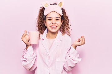 Beautiful kid girl with curly hair wearing sleep mask and pajamas holding coffee screaming proud, celebrating victory and success very excited with raised arm