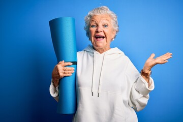 Senior beautiful sporty woman holding mat for yoga standing over isolated blue background very...