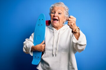 Senior beautiful sporty woman holding skate standing over isolated blue background screaming proud and celebrating victory and success very excited, cheering emotion - Powered by Adobe