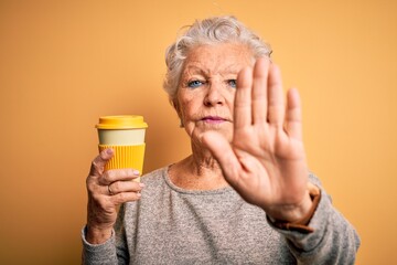 Senior beautiful woman drinking cup of coffee standing over isolated yellow background with open hand doing stop sign with serious and confident expression, defense gesture
