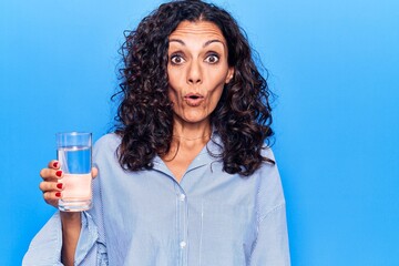 Middle age beautiful woman holding glass of water scared and amazed with open mouth for surprise,...