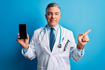 Middle age handsome grey-haired doctor man holding smartphone showing screen very happy pointing with hand and finger to the side