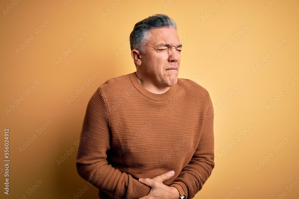 Wall mural middle age handsome grey-haired man wearing casual sweater over yellow background with hand on stoma