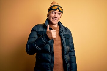Middle age handsome grey-haired skier man on vacation wearing ski goggles doing happy thumbs up gesture with hand. Approving expression looking at the camera showing success.